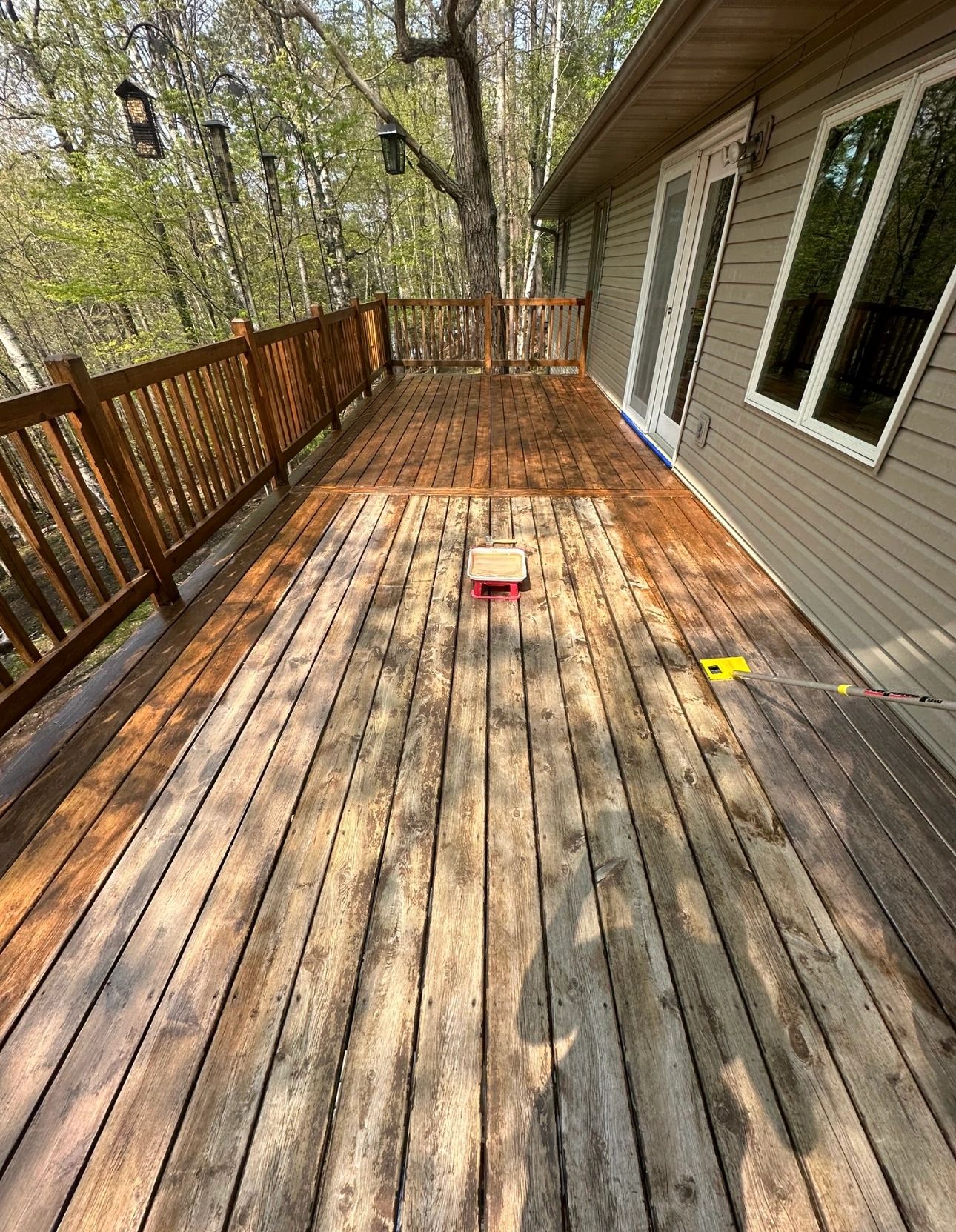 Wooden deck mid-stain beside a wooded house. Fresh cedar stain on the far boards and railing, weathered boards in the foreground. Photo from the Property Management Solutions site.